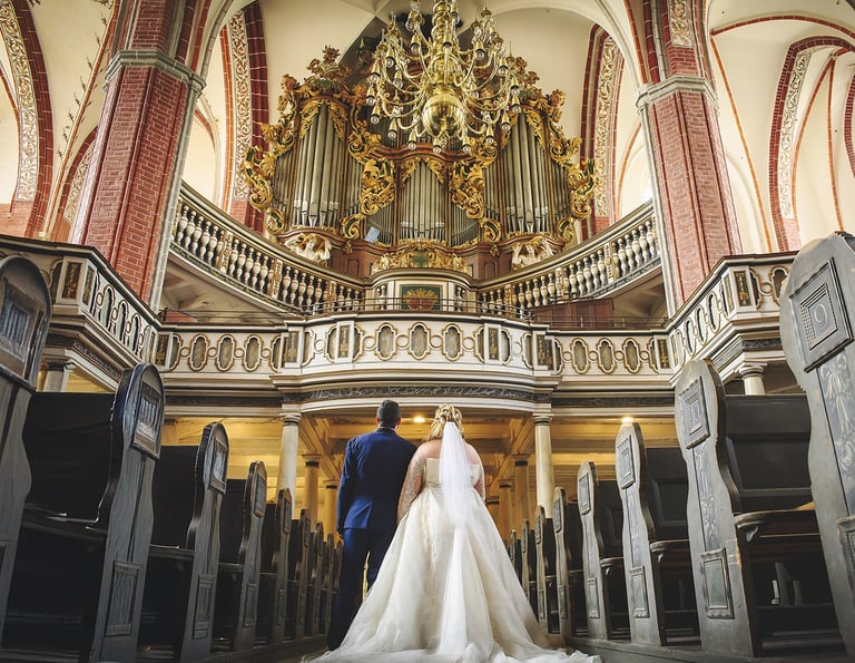 Ein Rückansicht-Foto eines Brautpaares, das in einer Kirche steht und auf eine große, verzierte Orgel blickt. Der Bräutigam 