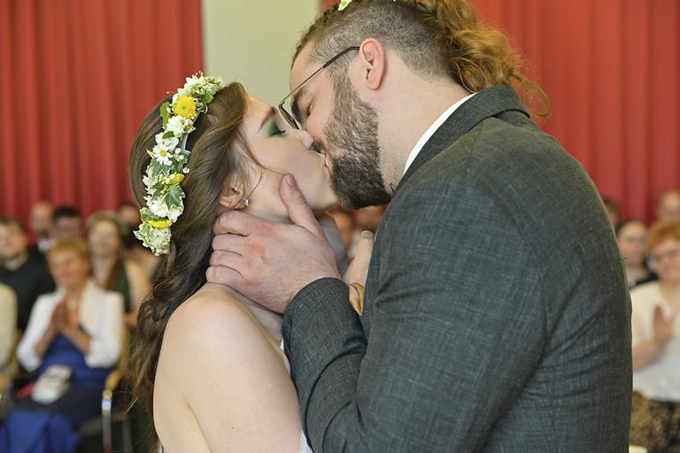 Ein Brautpaar küsst sich während der Trauung innig vor den applaudierenden Hochzeitsgästen. Die Braut trägt ein Blumenkranz i