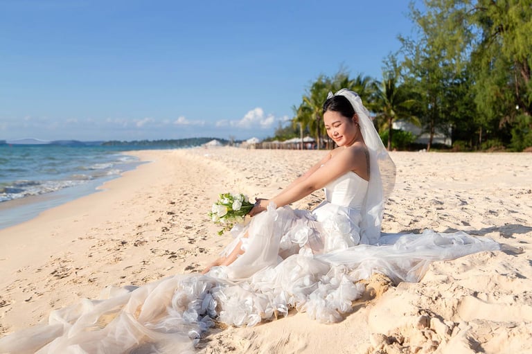 Bride in flowing gown sitting on quiet sandy beach