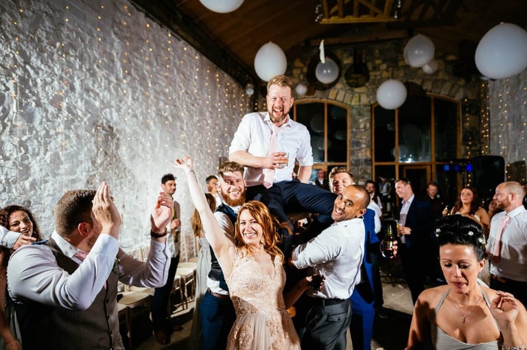 Wild party vibes at Rosedew Farm wedding as the groom is lifted high, guests cheering under fairy lights and balloons.