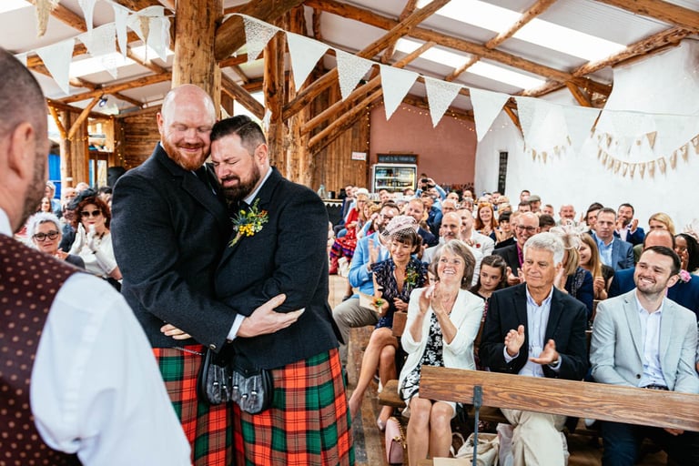Grooms embrace during wedding ceremony in rustic barn at South Wales venue Ceridwen, with guests clapping and cheering