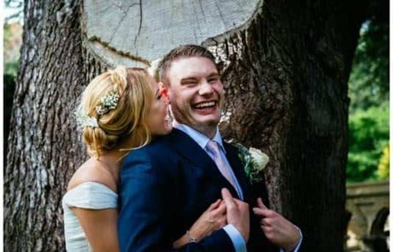 Bride and groom share laughter by a giant tree trunk, captured in a fun and natural wedding photography moment.