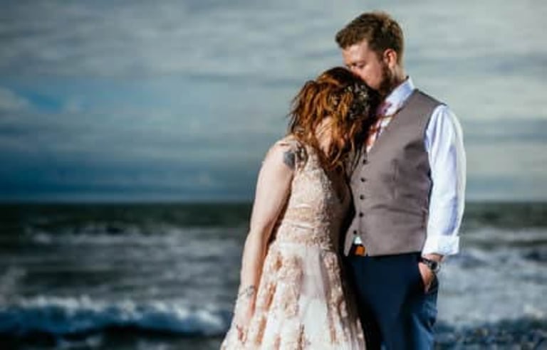 Newlyweds share a quiet embrace on Llantwit Major Beach, waves rolling in behind them under a moody sky.