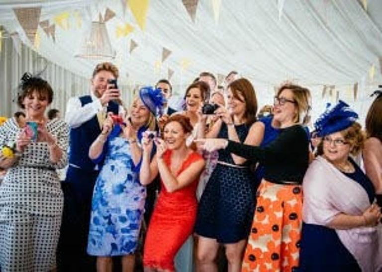 Wedding guests laughing, pointing and taking photos inside a marquee with bunting at a South Wales wedding, captured candidly