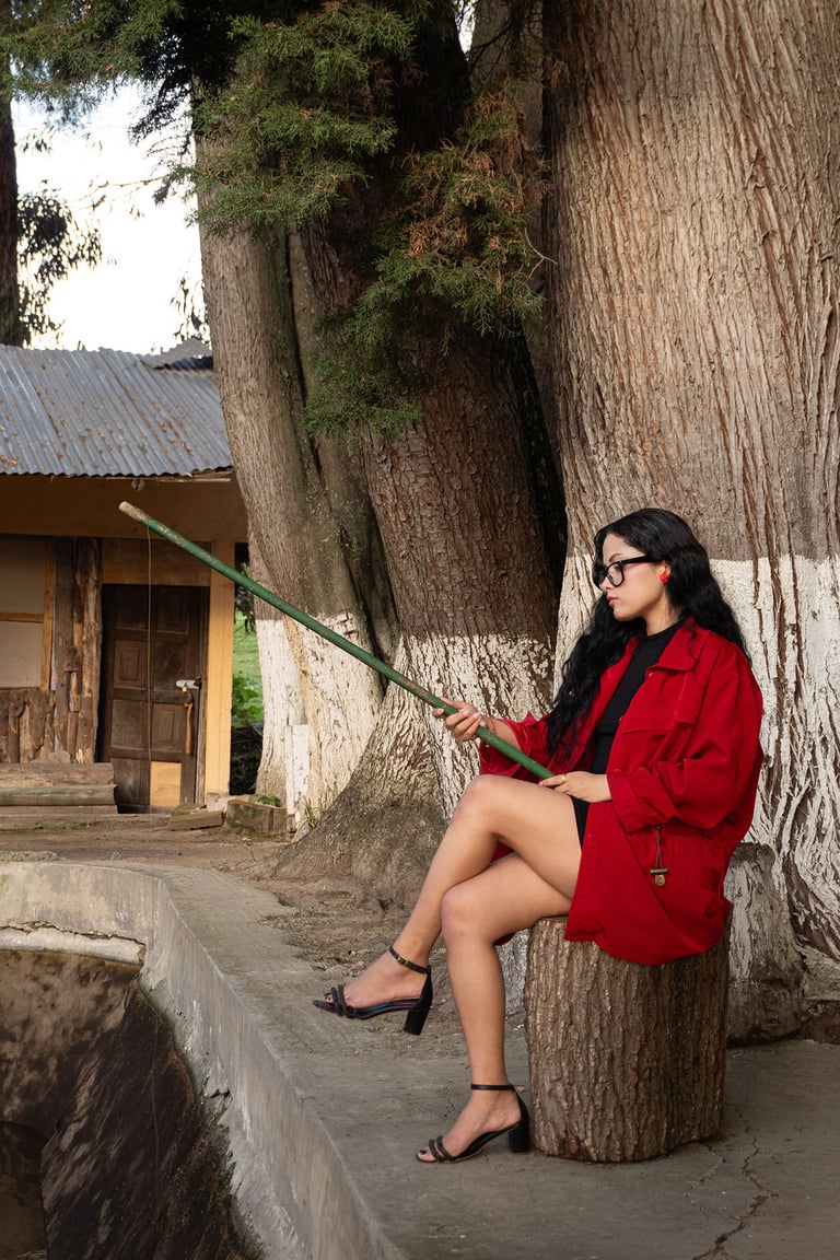 Mujer blanca de pelo negro con abrigo largo de color rojo, sentada en un tronco pescando 