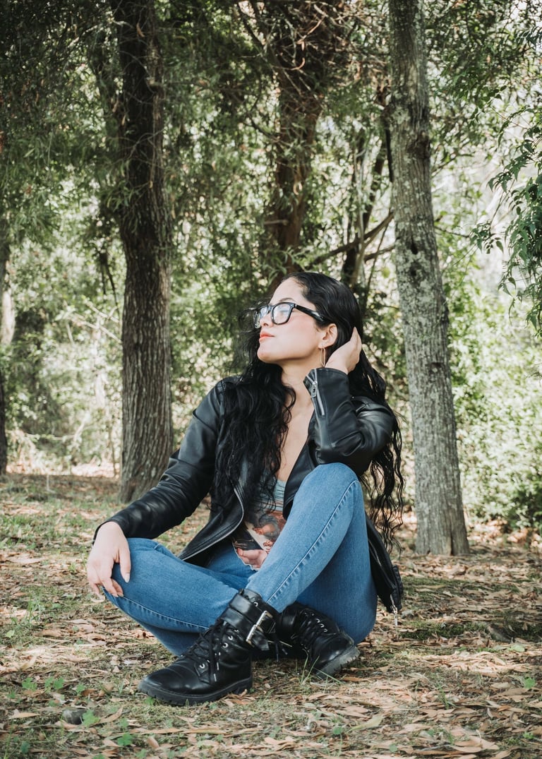 Mujer blanca de pelo negro sentada en medio del bosque con una chompa de cuero