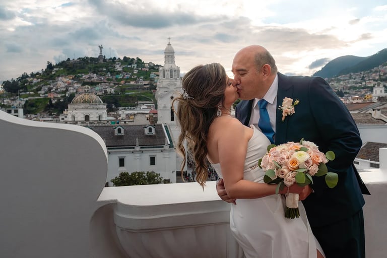 El novio besando a la novia mientras la agarra de la cintura en un terraza con vista al Panecillo en el centro histórico