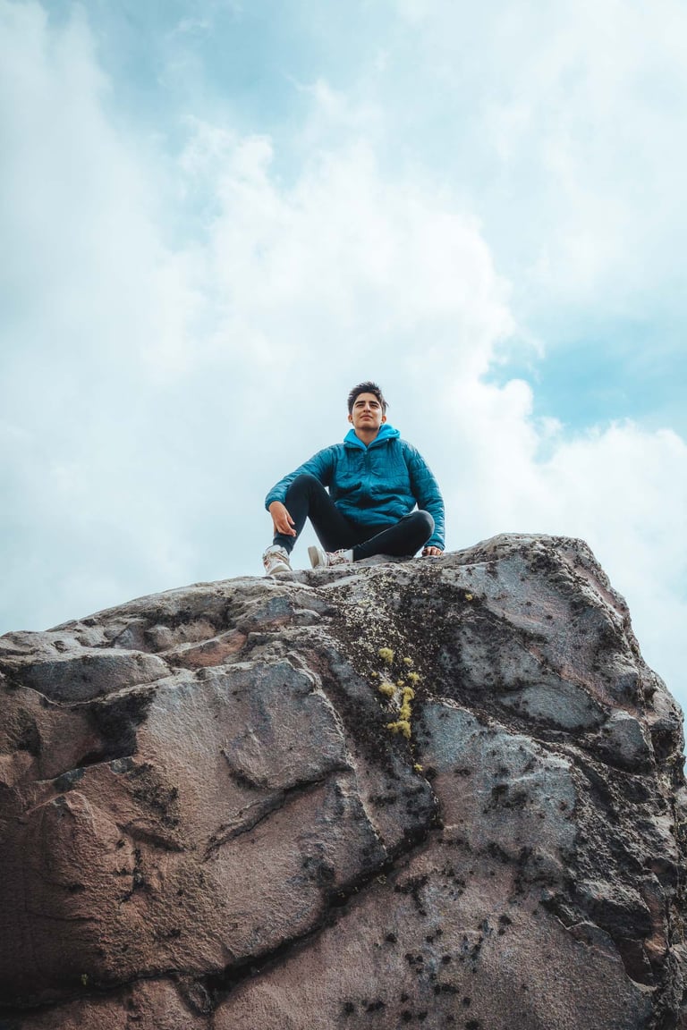 Retrato profesional creativo de aventura  de joven sentado en la cima de una roca con el cielo azul de fondo