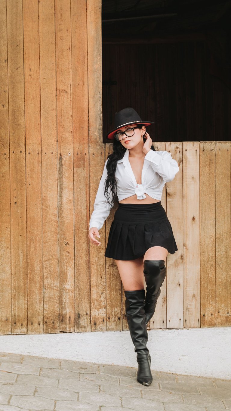 Mujer blanca de cabello negro con sombrero de vaquero, botas altas y camisa blanca en un establo