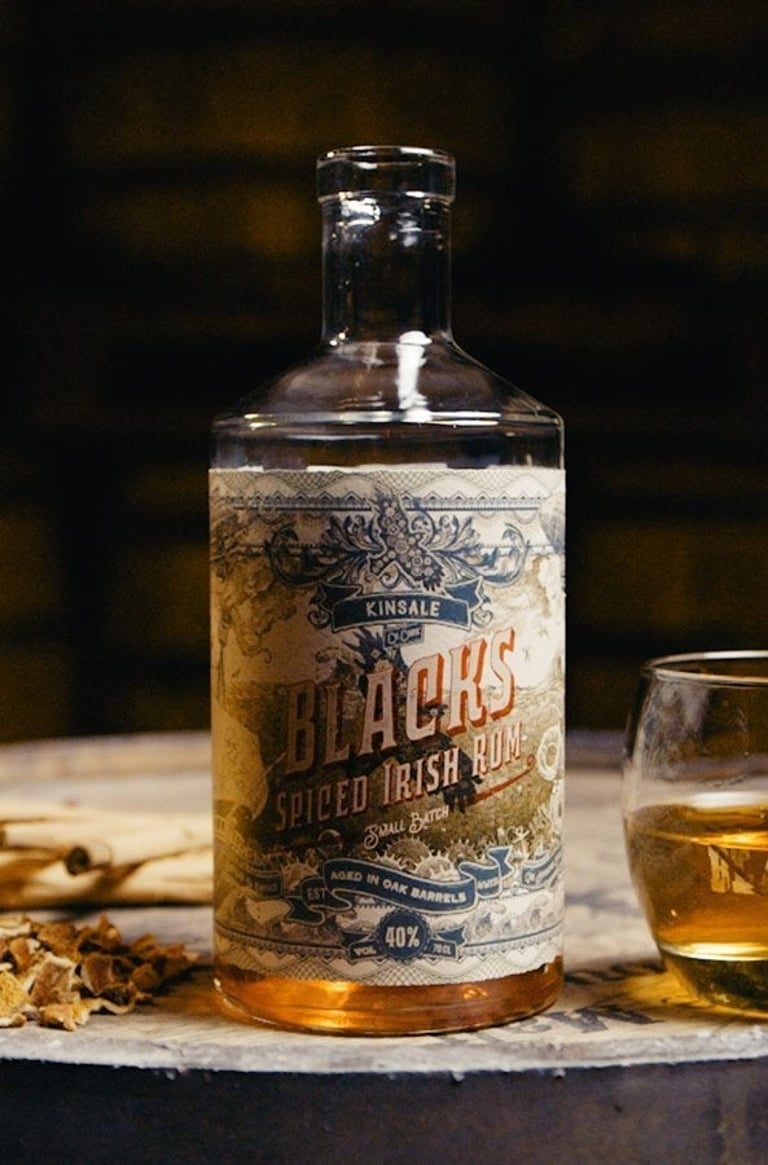 A bottle of Blacks Spiced Irish Rum on a wooden barrel with a glass of spirit and spices.