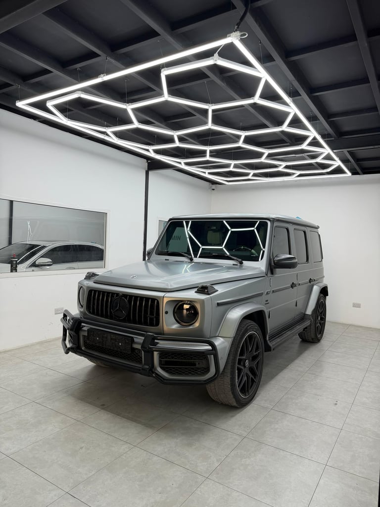 Matte grey Mercedes-Benz G-Class SUV parked under hexagonal LED garage lights in a luxury detailing studio.