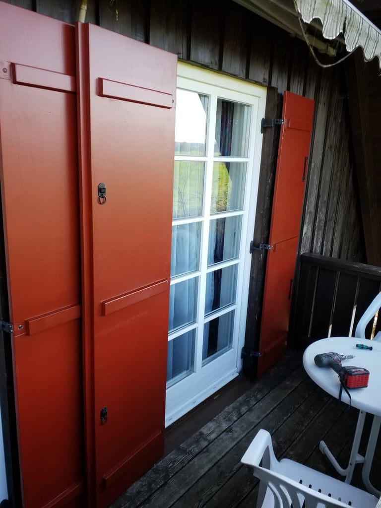 Weißes Holzfenster mit Sprossen und montierten roten Fensterläden an dunkler Holzfassade, Montage auf Balkon.