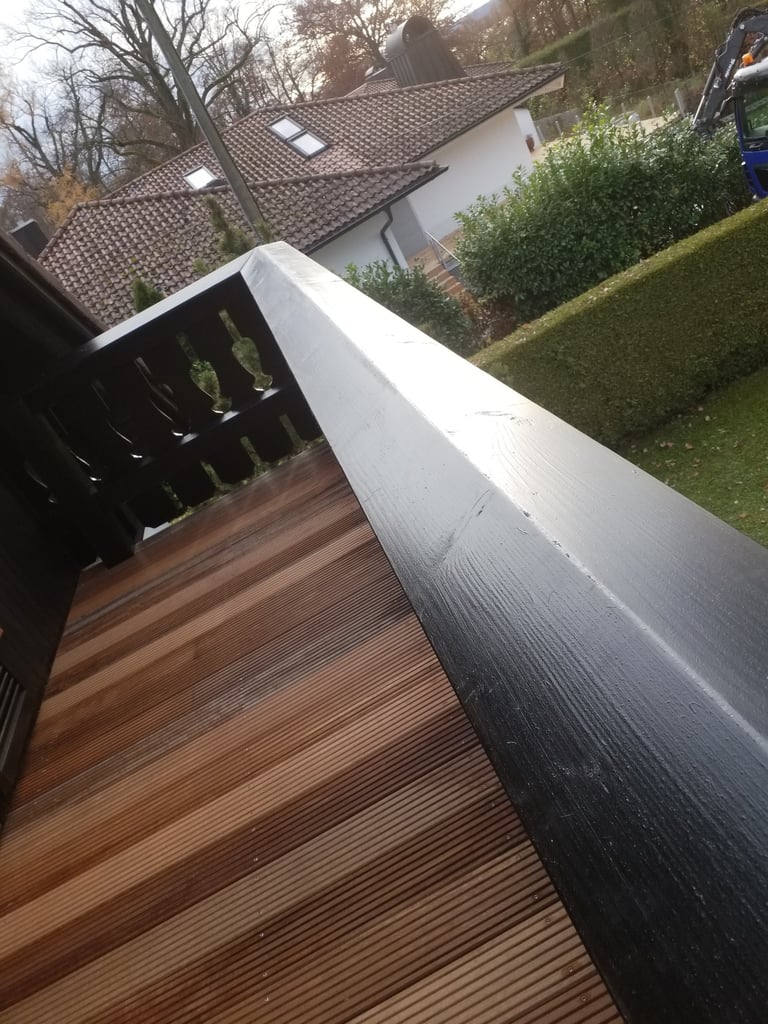 Nahaufnahme eines schwarzen Balkonhandlaufs mit Holzbohlen und Blick über den Garten auf ein Hausdach.