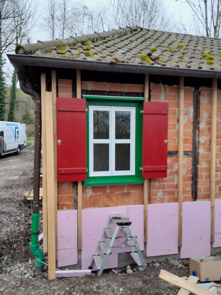 Eingebautes Holzfenster mit grünem Rahmen und roten Fensterläden an saniertem Ziegelmauerwerk.
