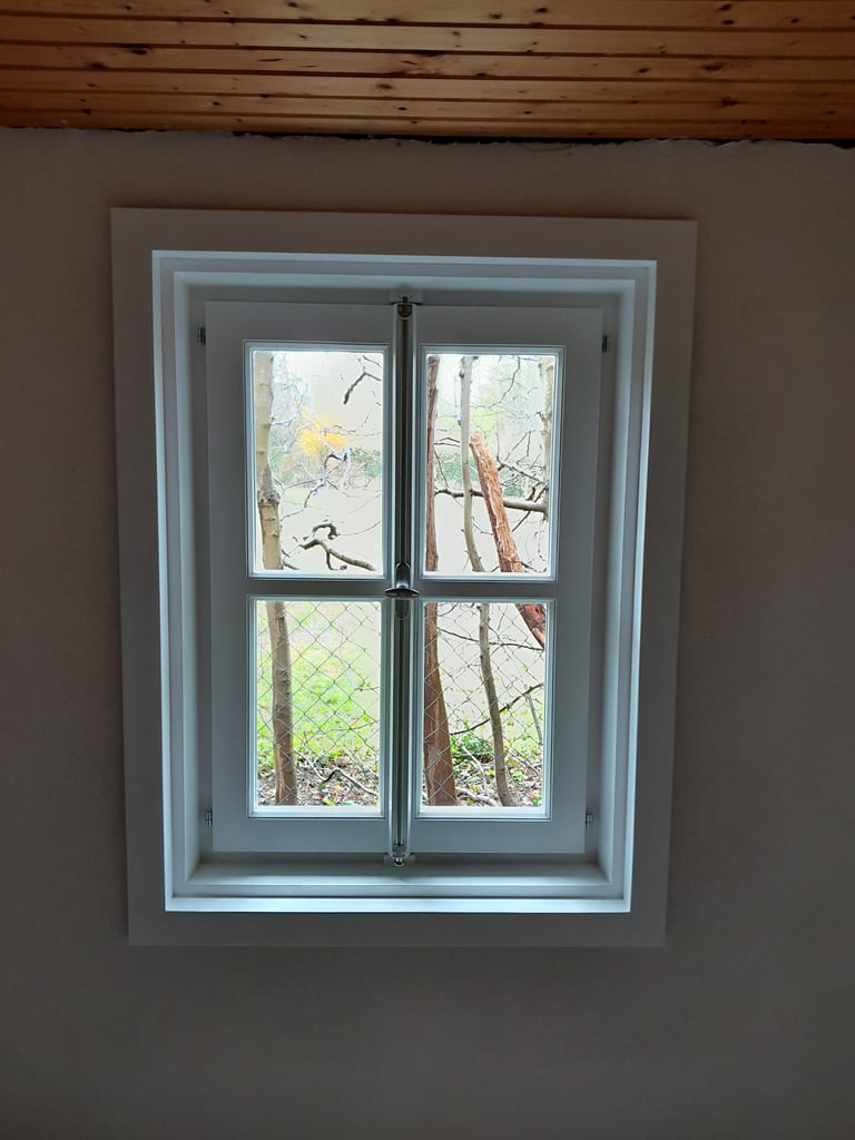 Weiß lackiertes Holzfenster mit Sprossen im Innenraum unter Holzdecke, Blick ins Grüne