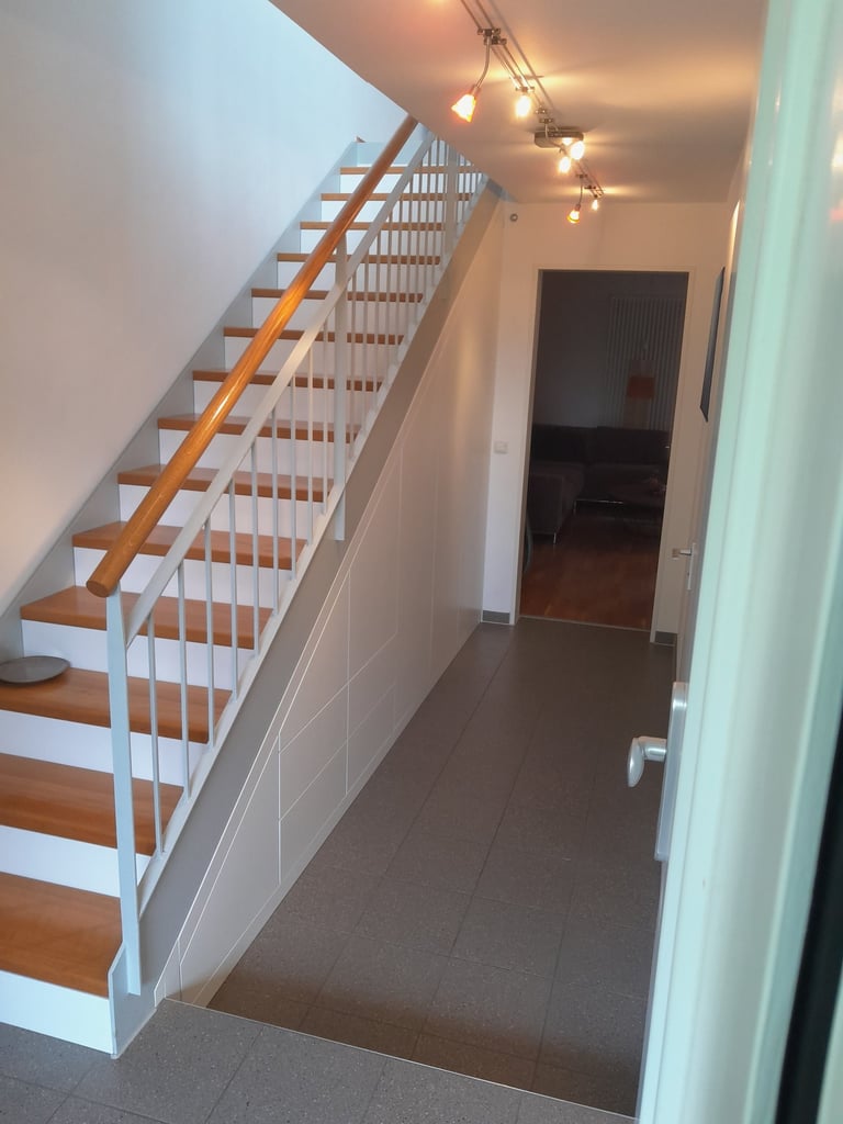 Heller Flur mit Holztreppe, weißem Geländer und Decken-Spots auf grauem Fliesenboden.
