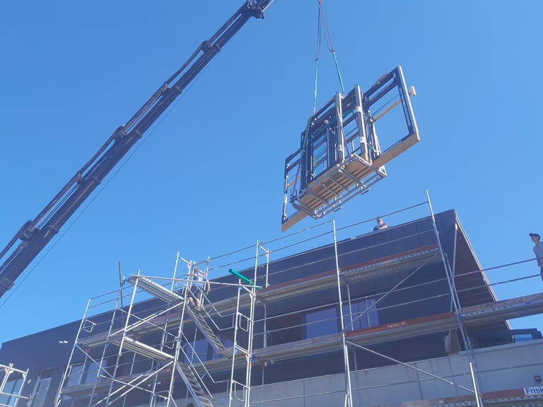 Kran hebt ein großes Fenster-/Fassadenelement auf ein Gerüst vor einem Gebäude.