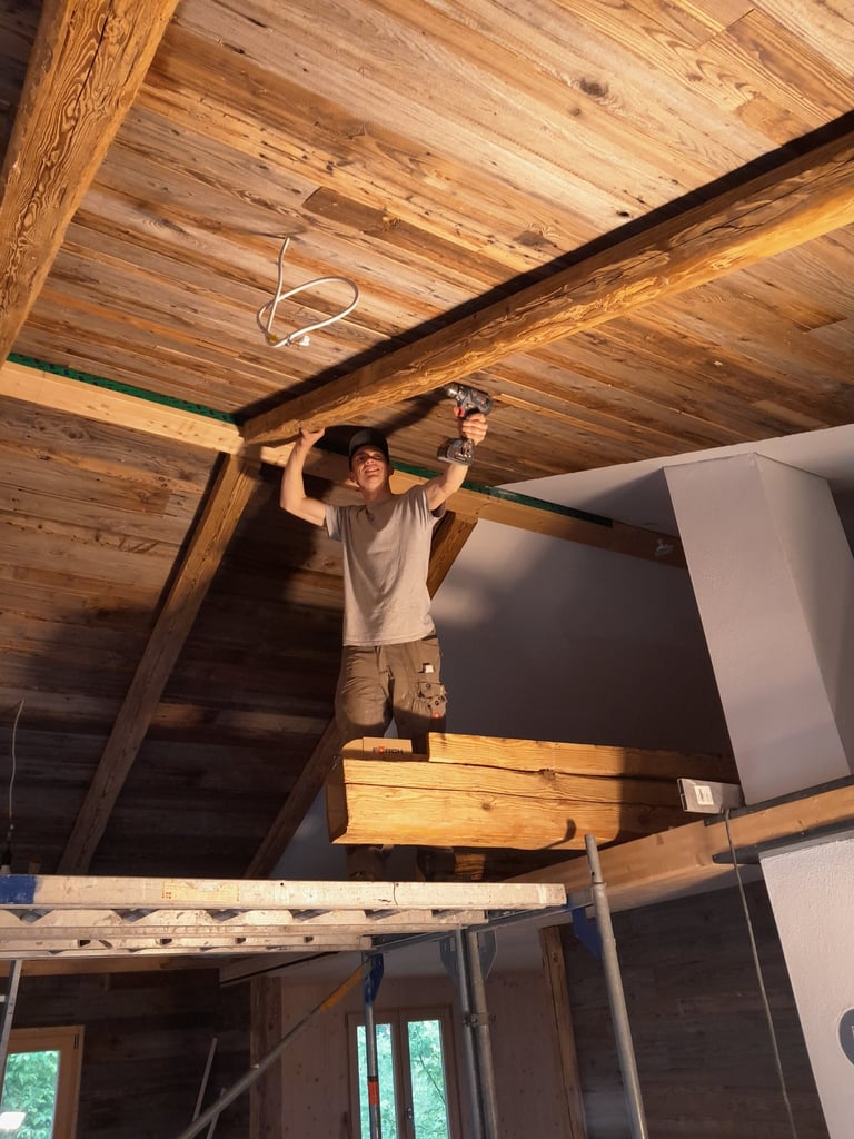 Schreiner montiert auf einem Gerüst eine rustikale Holzdecke mit Akkuschrauber.