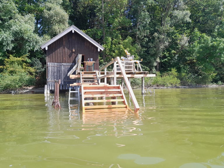 Holzhütte am Ufer mit im Bau befindlicher Holztreppe/Steg ins grüne Seewasser am Ammersee.