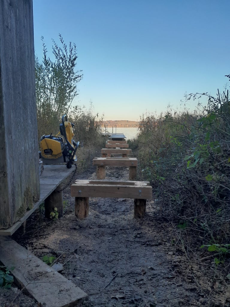 Holzunterkonstruktion für einen Steg führt durch Schilf zum Ammersee, daneben steht ein Rüttler.