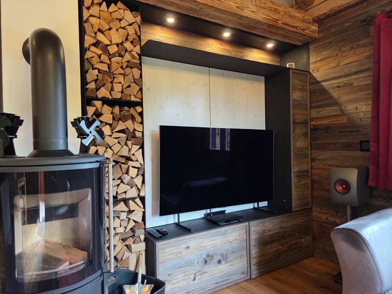 Wohnwand aus Altholz mit TV-Nische, integriertem Brennholzregal und Beleuchtung neben Kaminofen.