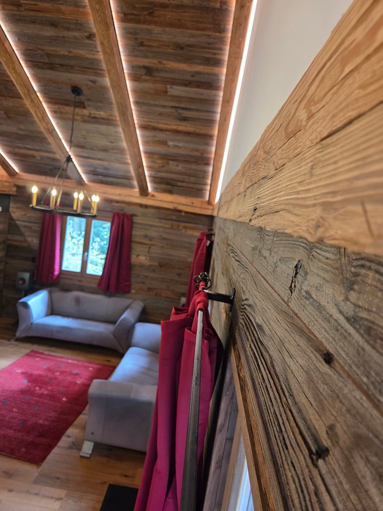 Gemütliches Wohnzimmer mit rustikaler Altholz-Wandverkleidung, Holzdecke mit LED-Lichtleisten und rotem Teppich.