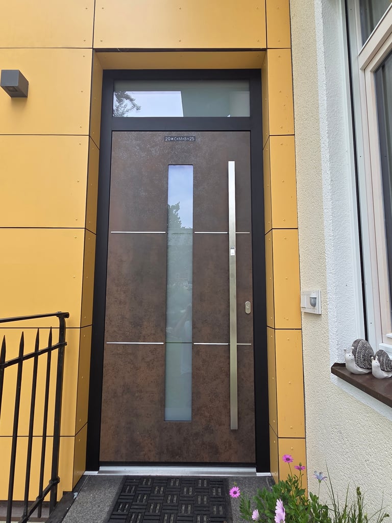Moderne Haustür mit vertikalem Glasausschnitt und Edelstahlgriff in dunkelbrauner Optik, eingebaut in gelbe Fassade,