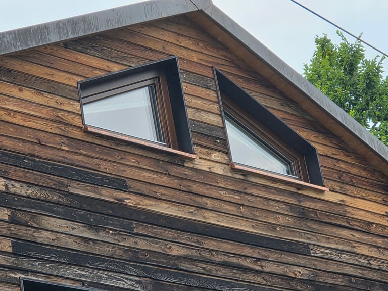 Holzfassade mit zwei schräg eingebauten Fenstern und dunklen Metallblenden, Kupfer-Fensterbänke am Giebel