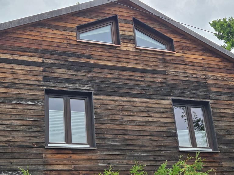 Hausfassade aus Holz mit vier Fenstern, oben zwei schräge Giebelfenster mit Metallblenden und Kupferbänken.