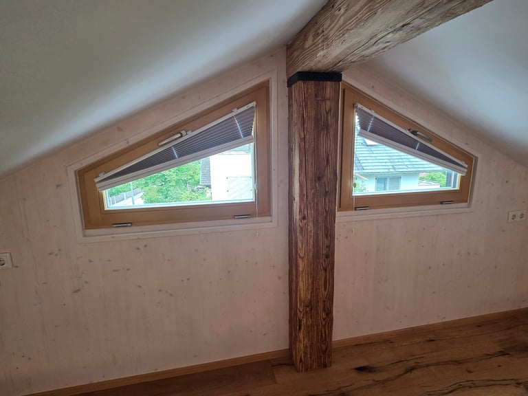 Innenausbau mit zwei schrägen Giebelfenstern aus Holz mit Plissees, sichtbarer Holzstütze und Holzboden im Dachraum.
