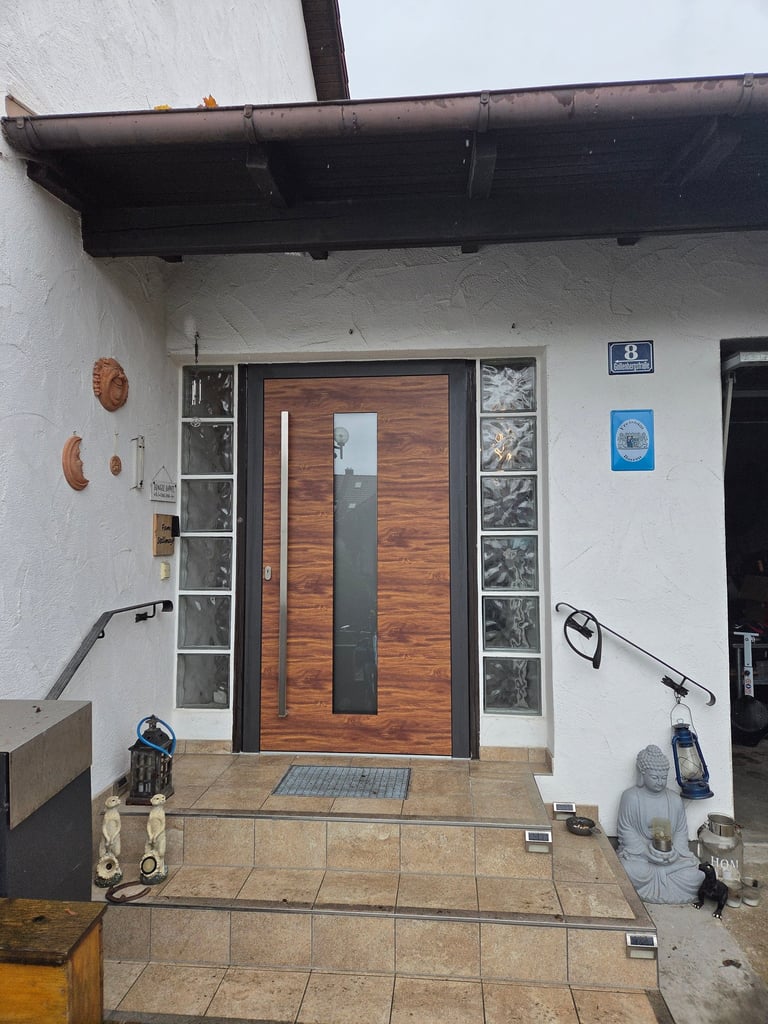 Eingebautes Haustür-Element in Holzoptik mit vertikalem Glasausschnitt und Edelstahlgriff, flankiert von Glasbausteinen.