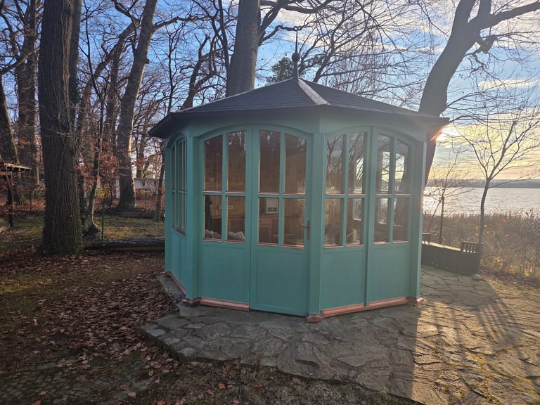 Grüner, verglaster Holzpavillon mit spitzem Dach und Kupferdetails auf einer Steinterrasse am Seeufer, umgeben von Bäumen.
