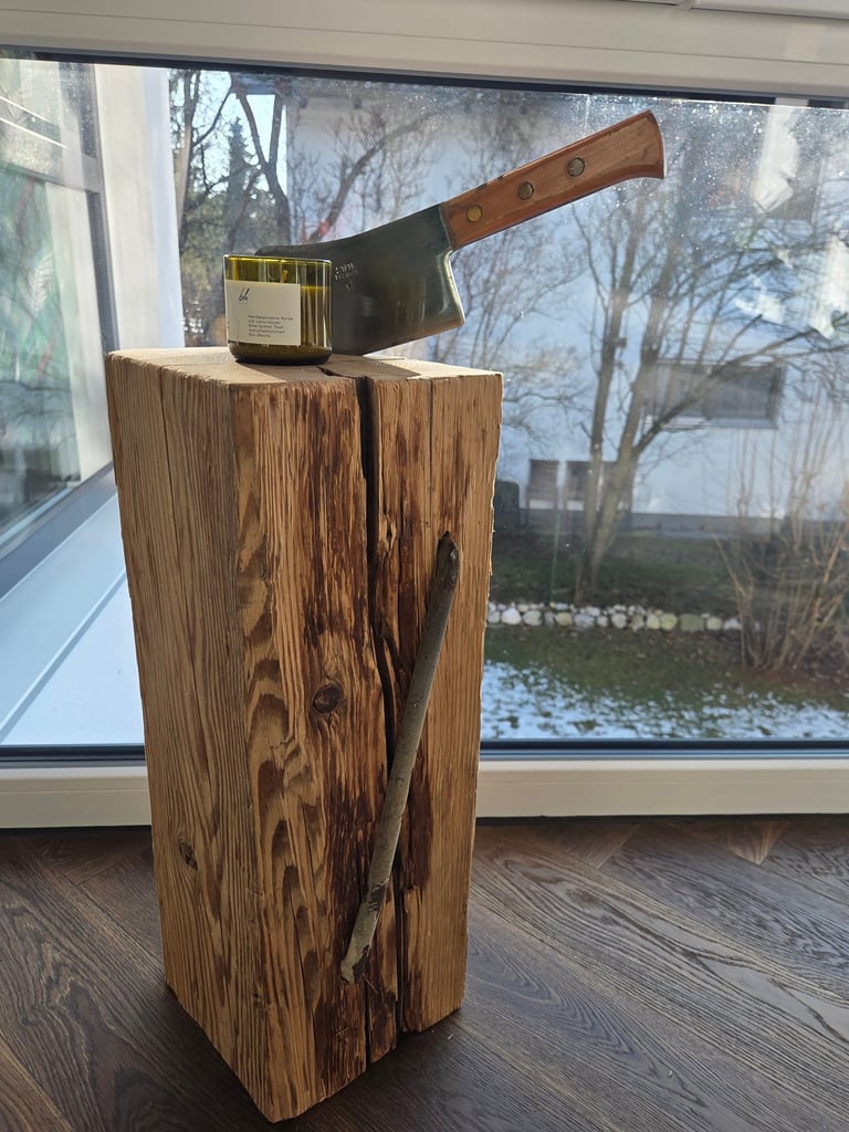 Rustikaler Holzblock als Deko-Objekt mit eingelassenem Eisenwerkzeug, darauf Beil mit Holzgriff und Kerze am Fenster.
