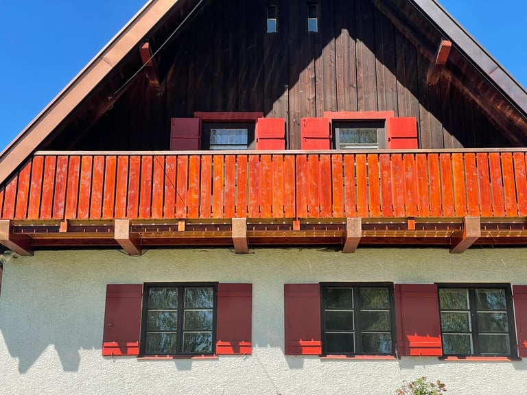 Traditioneller bayerischer Holzbalkon mit rotbraunen Sichtschutz-Latten und passenden Fensterläden an einem Wohnhaus.