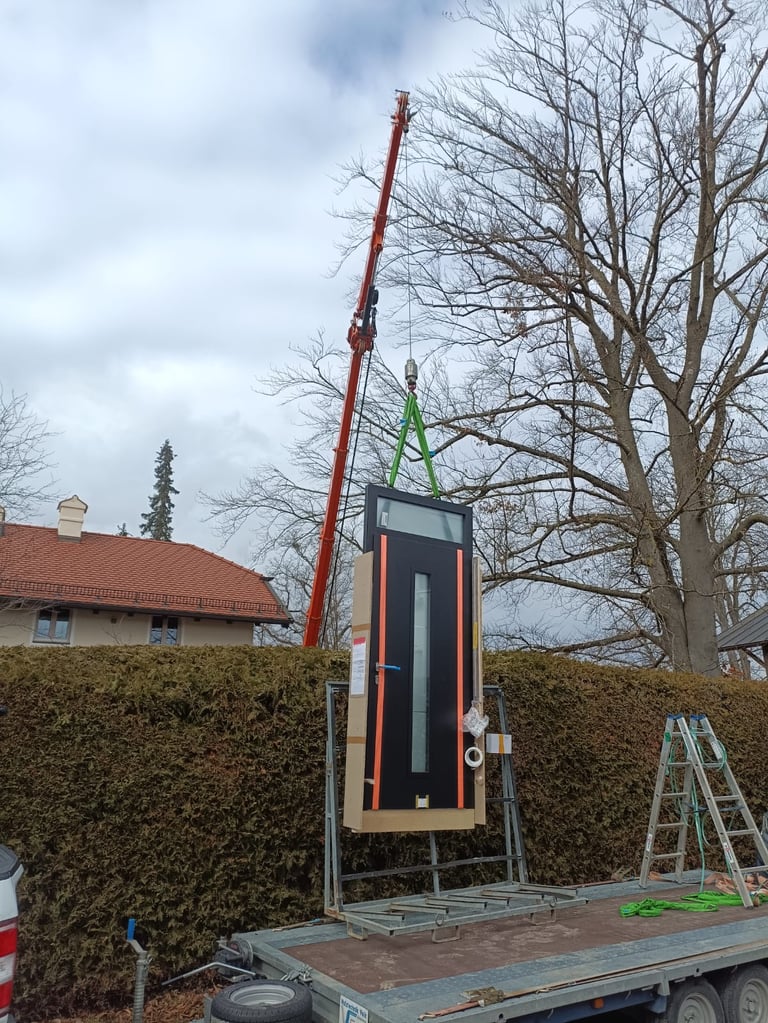 Kran hebt ein modernes Haustür-Element mit Glasausschnitt auf Anhänger zur Montage, Leiter und Baustellenumfeld