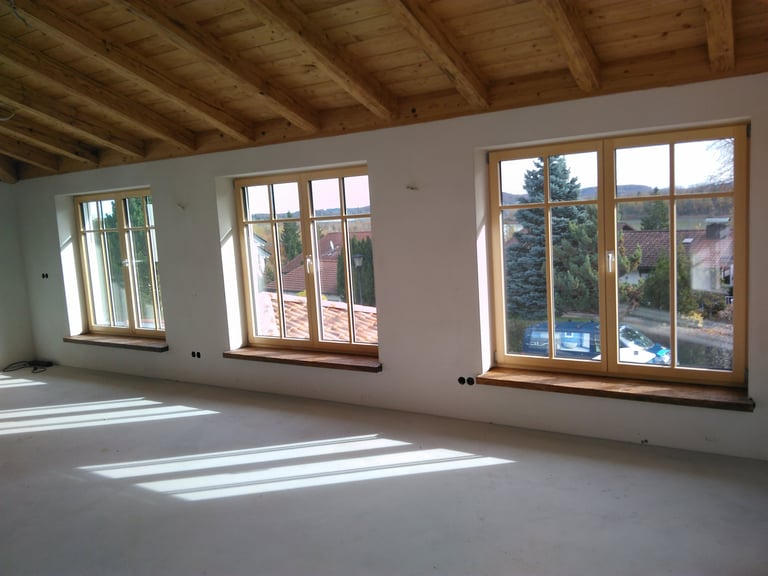 Heller Raum mit Holzdecke und drei Fenstern, durch die Sonnenlicht auf den Boden fällt.