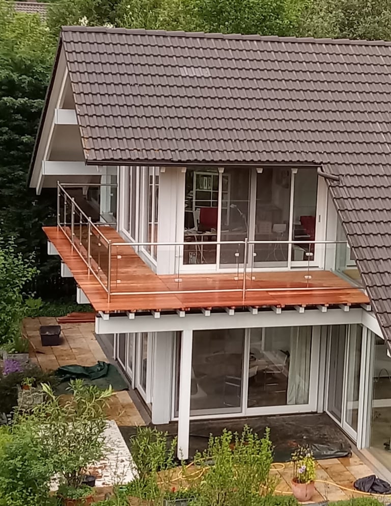 Modernes Haus mit großer Holzterrasse und Glasgeländer im Obergeschoss.