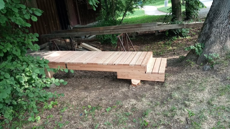 Holzpodest am Ammersee mit kleiner Stufe unter Bäumen neben einer Scheune im Grünen.