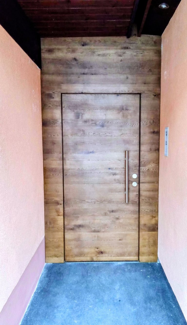 Holztür in einer holzverkleideten Wandnische mit langen Griff, flankiert von orangefarbenen Wänden.