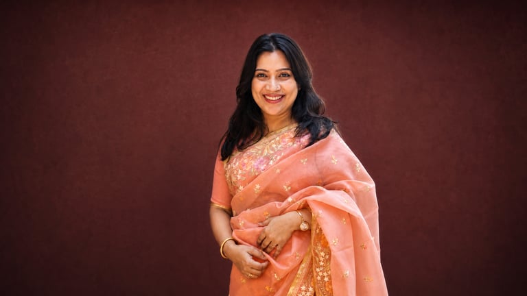 Smiling woman in an elegant peach silk saree with floral embroidery against a dark red wall.