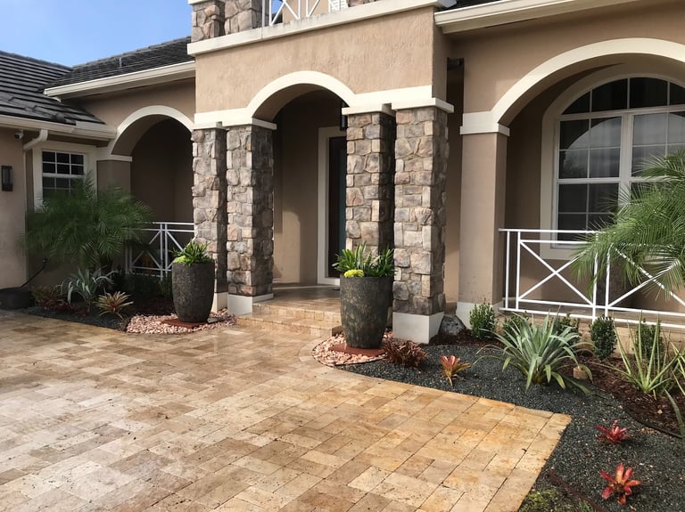 Luxury home entrance with stone pillars, travertine paver driveway, and tropical landscaping.