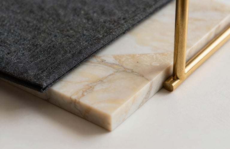 Close-up of premium interior design materials: a sample of polished champagne marble, a charcoal fabric swatch, and a gold metal furnishing detail on an off-white surface.