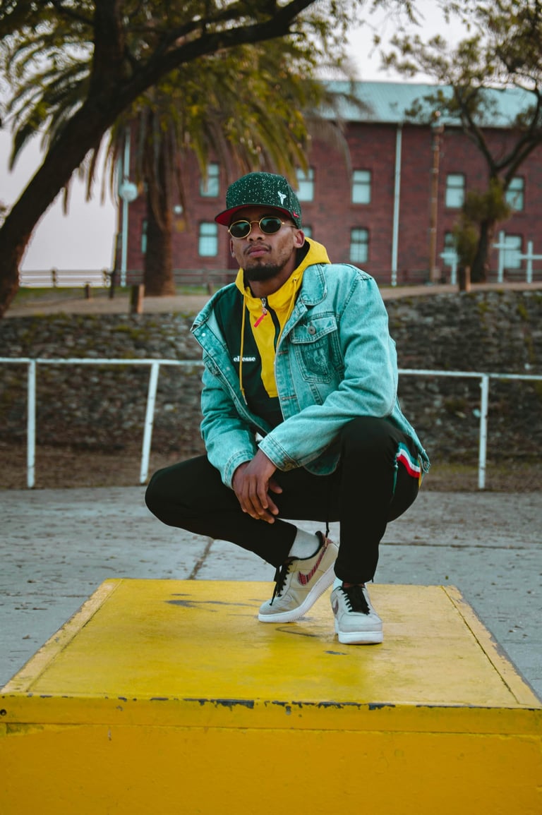 un homme accroupi qui porte une casquette, une veste en jeans un sweat jaune et une paire de nike