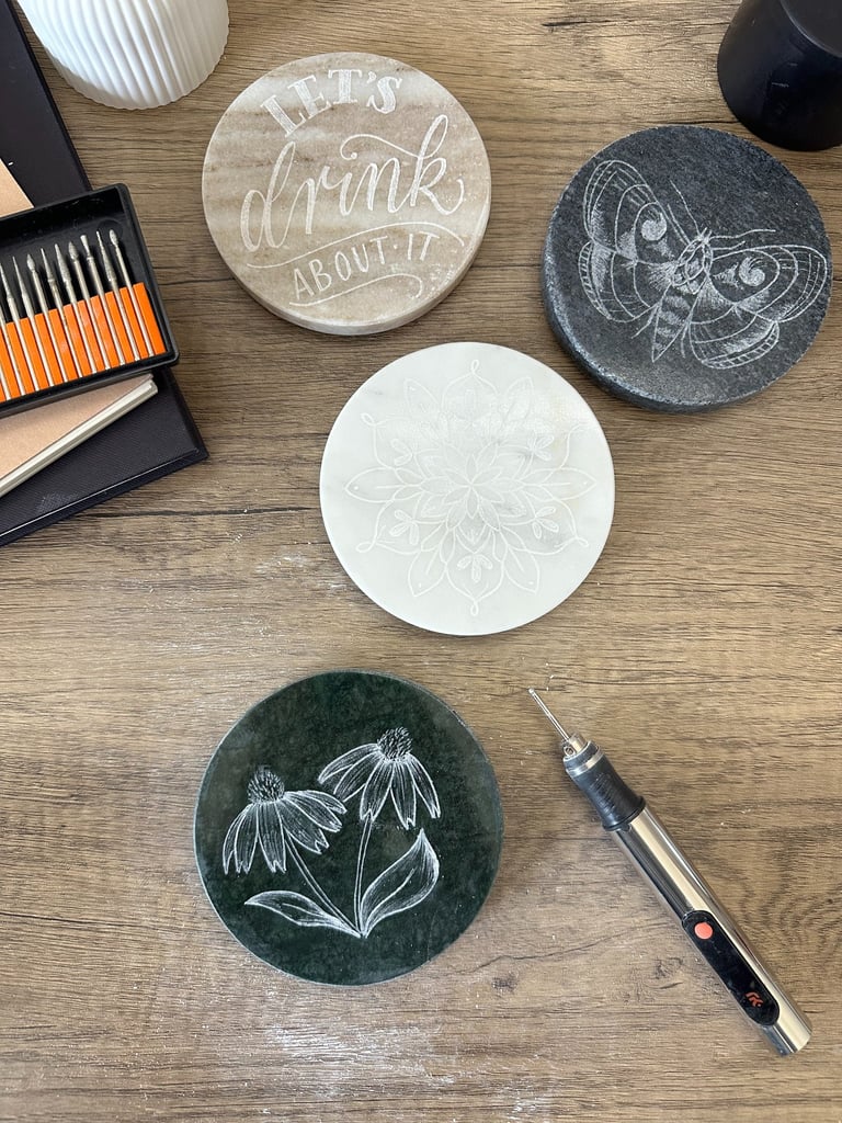 Engraved stone coasters with floral and moth designs next to a rotary engraving tool.
