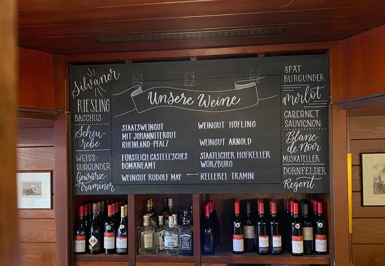 Handwritten chalk wine menu listing Silvaner, Riesling, and Merlot above a shelf of bottled German wines.