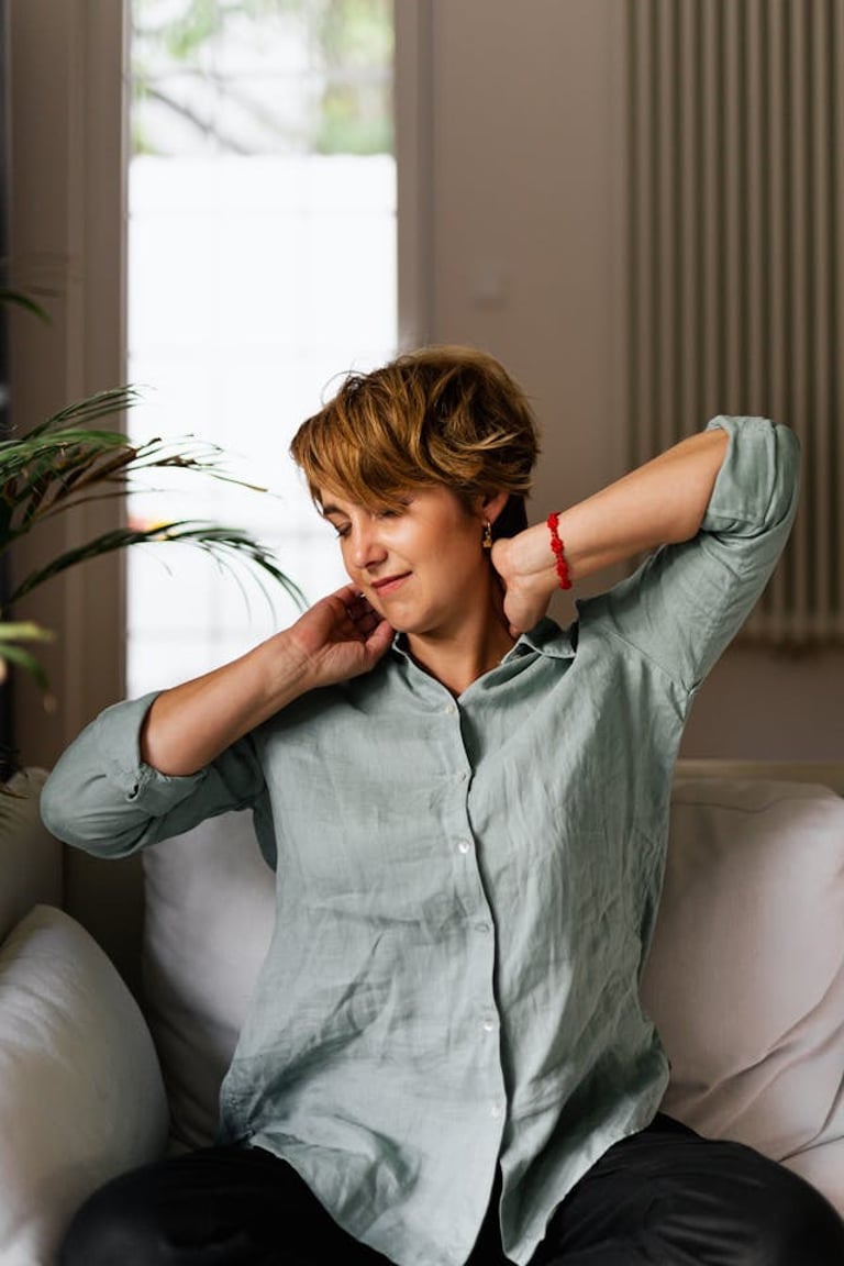 A woman in a green linen shirt relaxes at home while massaging her neck to relieve tension.