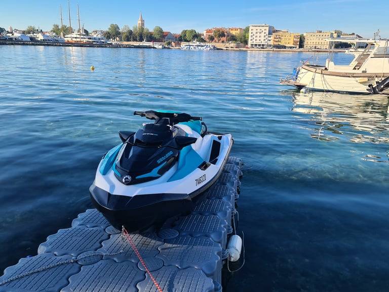 A blue and white Sea-Doo jet ski parked on a floating dock in a sunny European harbor.