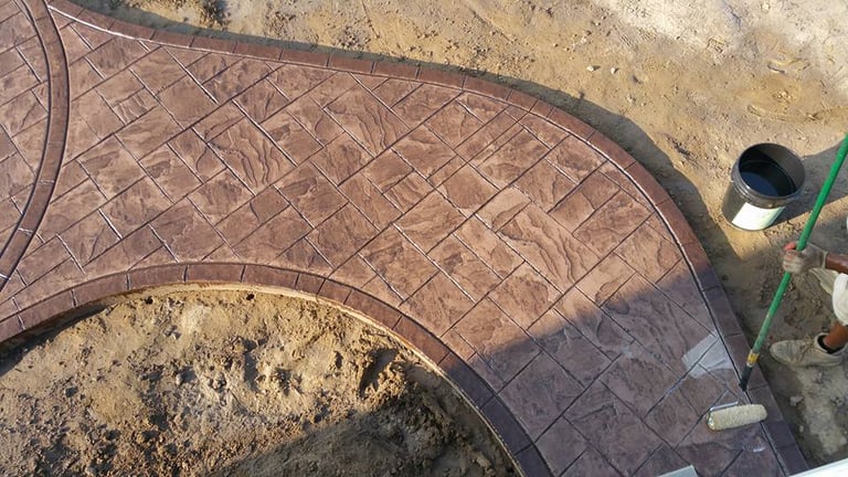A worker applying a clear sealant to a curved brown stamped concrete walkway with a slate stone pattern.