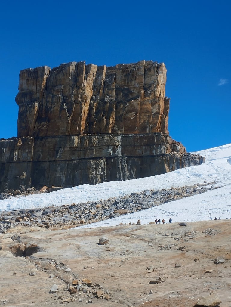 Hikers trekking past a massive rock formation and a snowy glacier under a clear blue sky.