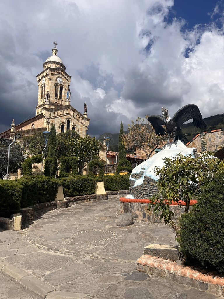 Historic stone church with a bell tower and an Andean condor statue in a scenic Colombian town square.